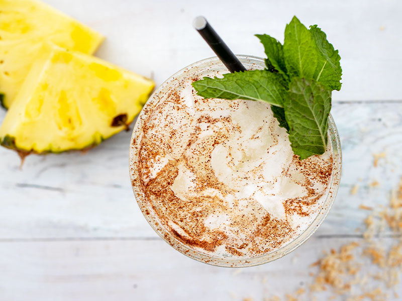 Aerial shot of a Tocaya drink, garnished with mint and cinnamon, with pineapple on the table in the upper left hand corner of the photo.