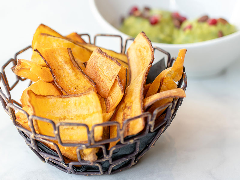 Close up image of a plantain chips in a metal bowl, with guacamole blurred in the background.
