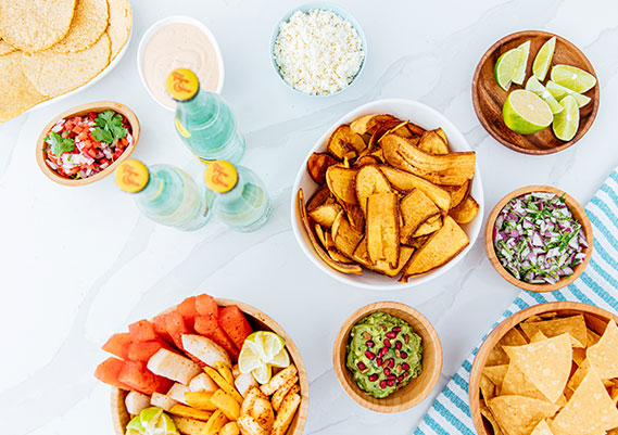 Aerial view of plantain chips, guacamole, chips, salsa, clear soda and the barrio fruit bowl sitting on a white marble table.