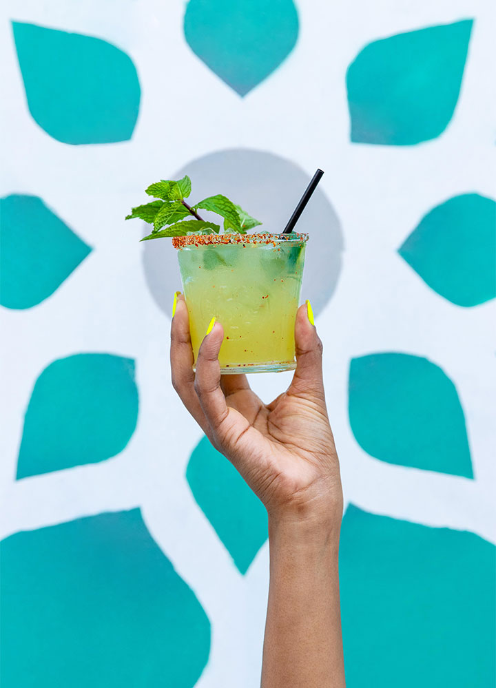 A woman with yellow nail polish holding up a yellow margarita with a black straw and garnished with mint in front of a blue and white lotus designed backdrop.