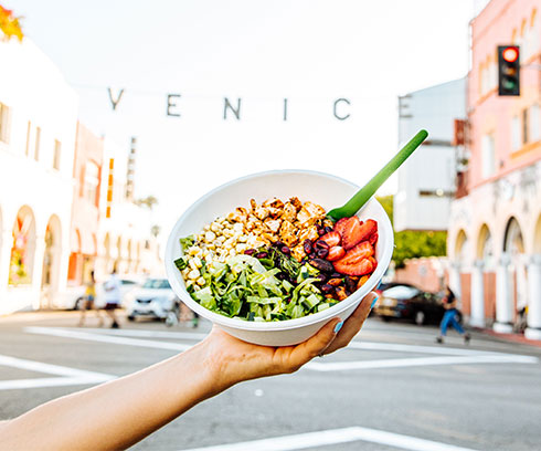 Shot of an arm holding up a Tocaya salad in downtown Venice with the Venice sign stretched out across the street in the background.